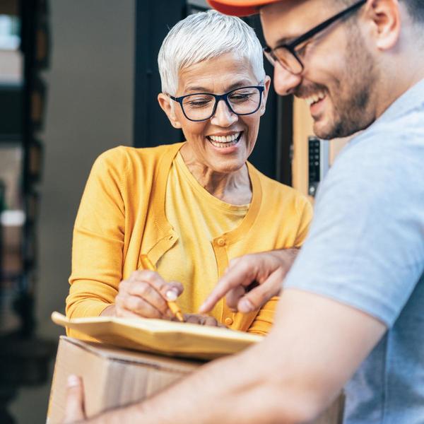 Foto de una señora sonriente con gafas y jersey amarillo firmando la entrega de un paquete a un mensajero con gorra roja y camiseta gris
