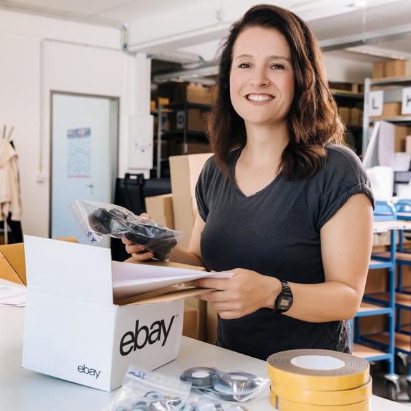 Foto de mujer joven con melena castaña sonriendo a cámara y empaquetando un artículo para su envío en una caja con el logo de eBay