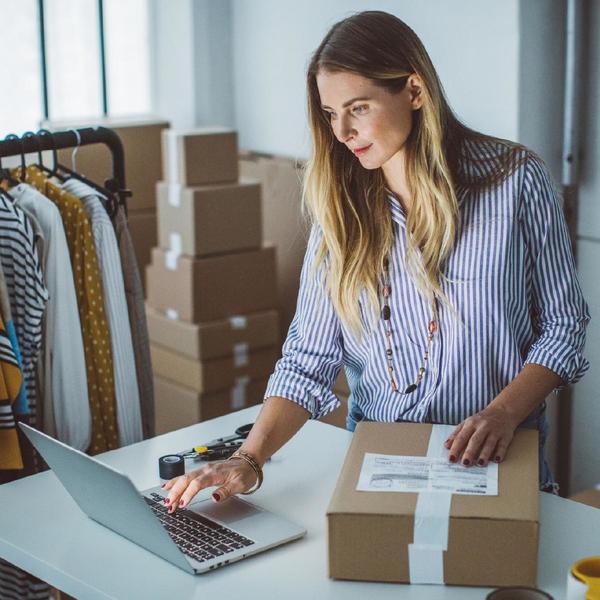 Woman looking at laptop with one hand on a package and other packages and a rack of clothes behind her