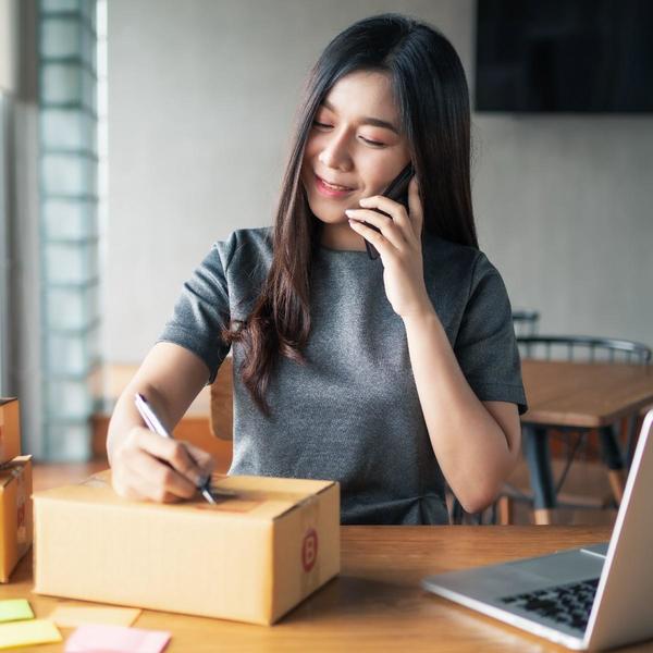 Woman on phone, writing on package with a laptop open beside her