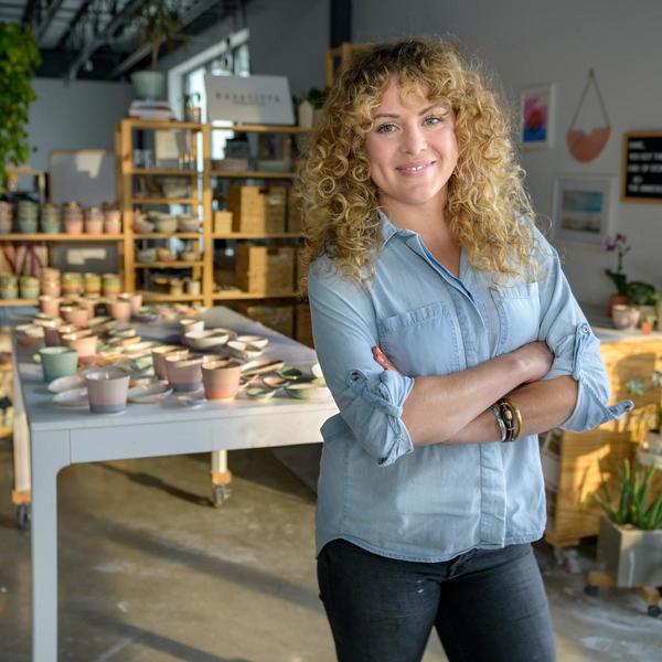 Real seller photo: blonde, curly-haired woman standing in front of her pottery with crossed arms