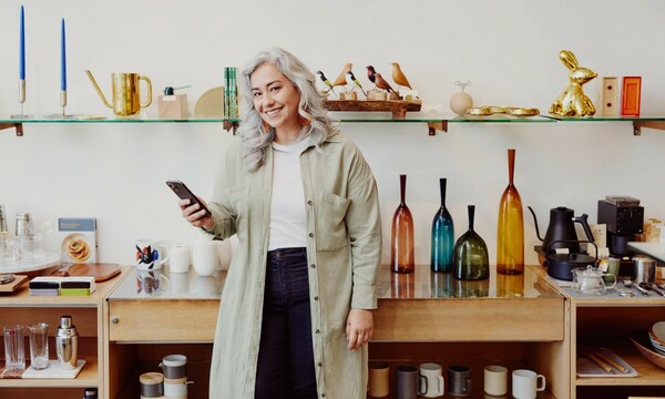 Woman smiling and holding a phone in front of a shelf of ceramics and vases.