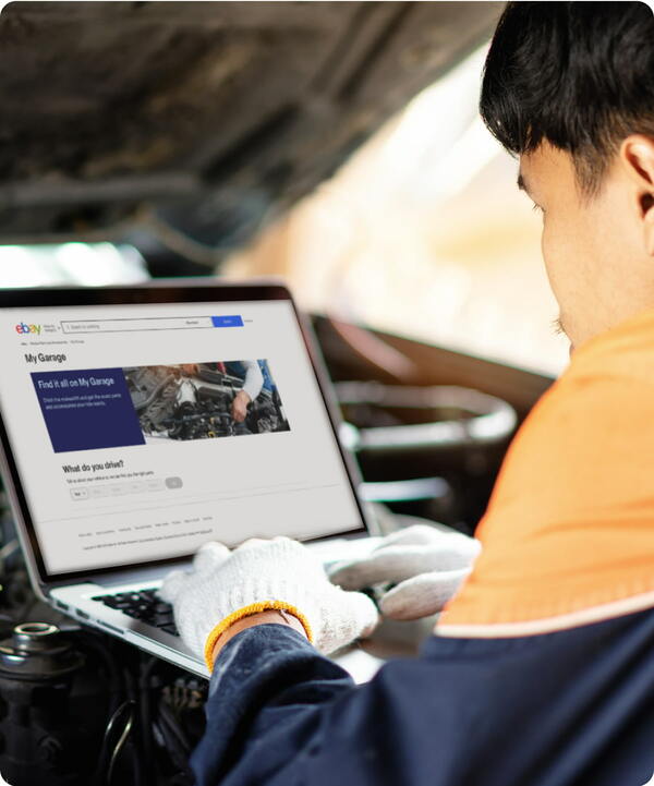 A person wearing an orange and navy jacket and work-gloves is leaning toward their laptop, which is sitting on the engine under a popped hood of a car. On the laptop screen, the eBay Motors ”My Garage“ page is open.