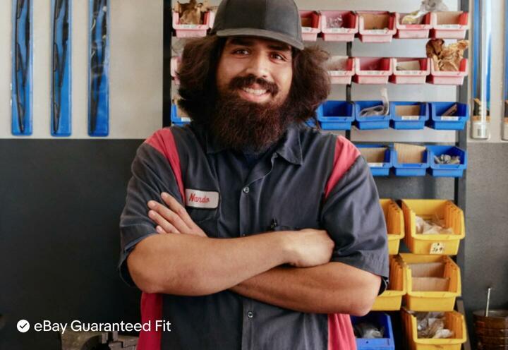 Nando, a mechanic, smiles at the camera with his arms folded around his chest, wearing a gray short-sleeve, work-shirt with red stripes on either side. Behind him, is a rack of red, blue, and yellow tool bins, a black and gray tool cabinet with cardboard boxes and a red rear-light stacked on top. Packaged windshield wipers are hung up on the empty of the wall. eBay Guaranteed Fit is shown on the bottom left of the image.
