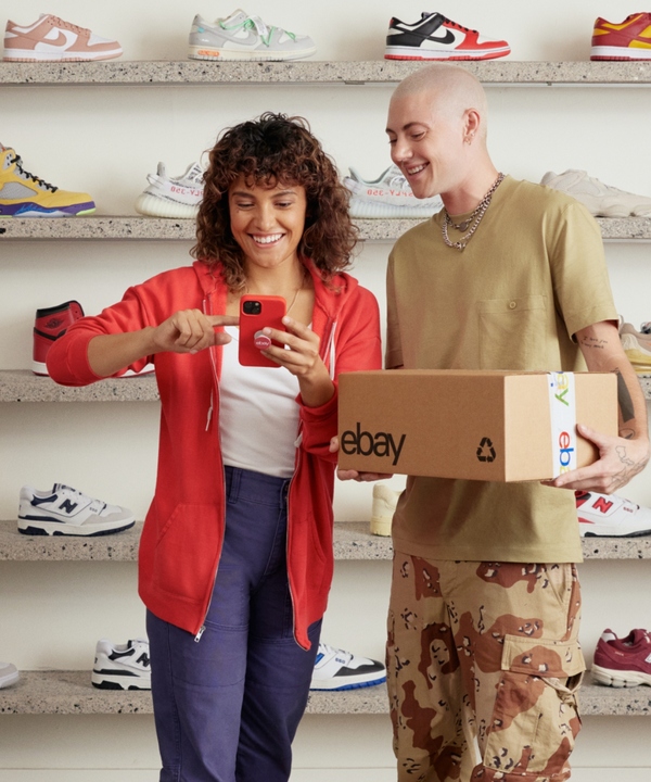 A lady wearing red sweater is showing something on her red phone to a man holding an eBay package. They're standing in front of a wall of sneakers.