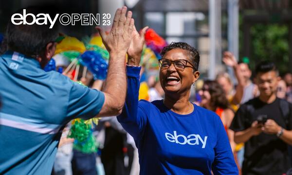 A woman wearing a blue eBay shirt is smiling and high-fiving someone at a parade. Behind her, a crowd of people are holding colorful pom-poms. The eBay Open 2023 word mark sits in the top-left corner.