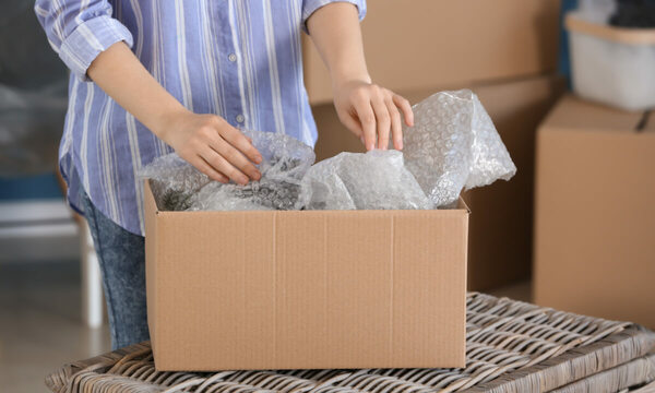 An eBay reseller adding bubble wrap within a shipping box on a wicker table with more packages stacked in the background.