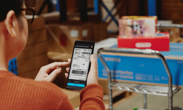Reseller viewing shipping details through the eBay app with the packaged product in the background of a warehouse.