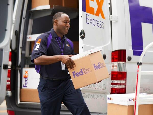 A FedEx delivery man grabbing 2 packages from the delivery van and stacking the boxes on a trolley.