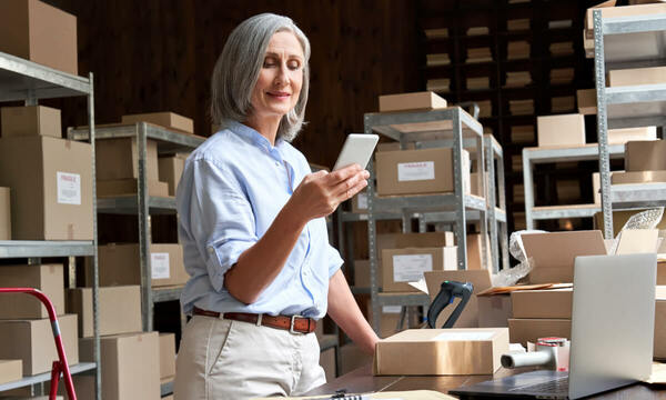 A woman standing in a warehouse surrounded by shelves of packages looking at a tablet with incoming order. She is leaning on a desk with packages and shipping materials on top.