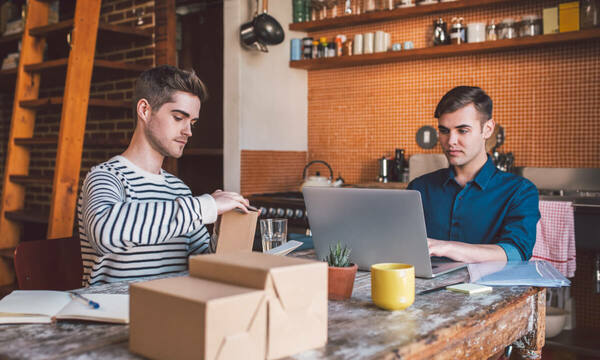 Two eBay sellers sitting at a table preparing boxes for delivery and managing orders on their laptop.