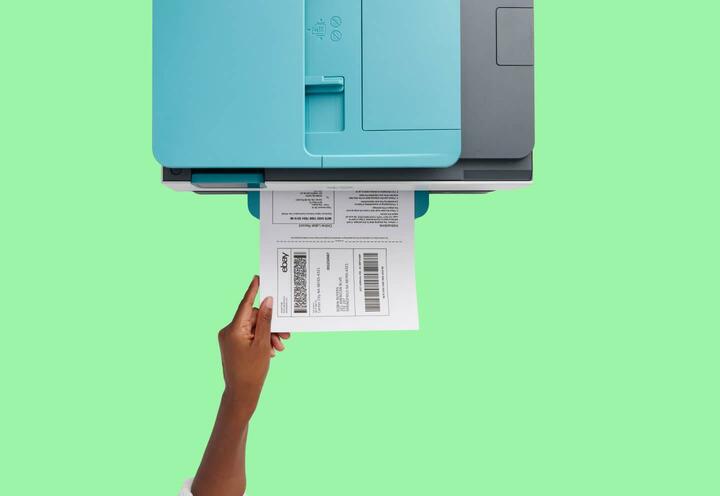 A woman grabbing an eBay shipping label from the tray of a printer with a green backdrop.