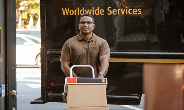 A UPS delivery man wheeling a stack of three packages through the warehouse on a dolly with the brown and yellow UPS van in the background.