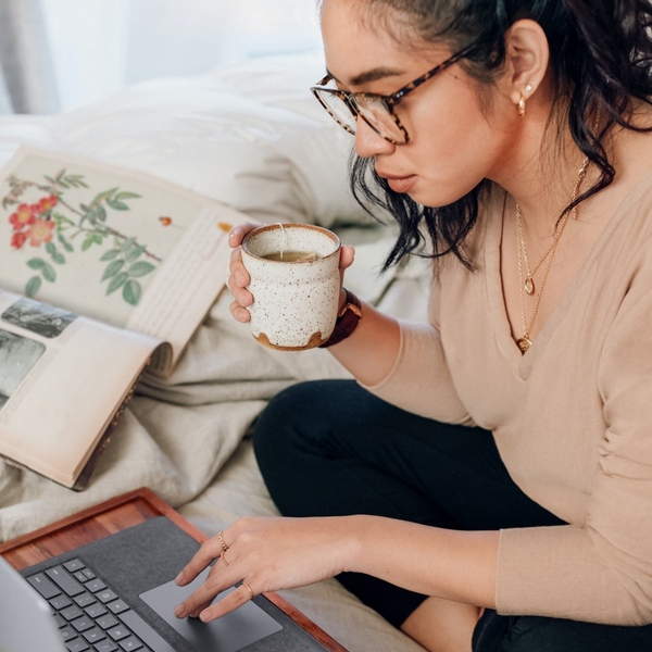 Frau mit Kaffeetasse in der Hand blickt auf den Bildschirm ihres Laptops.