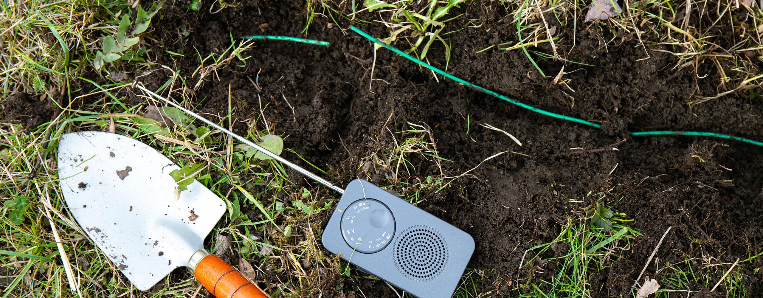 Eine Gartenschaufel und ein mobiles AM-Radio liegen neben einem ausgegrabenen Mähroboter-Kabel mit Kabelbruch.