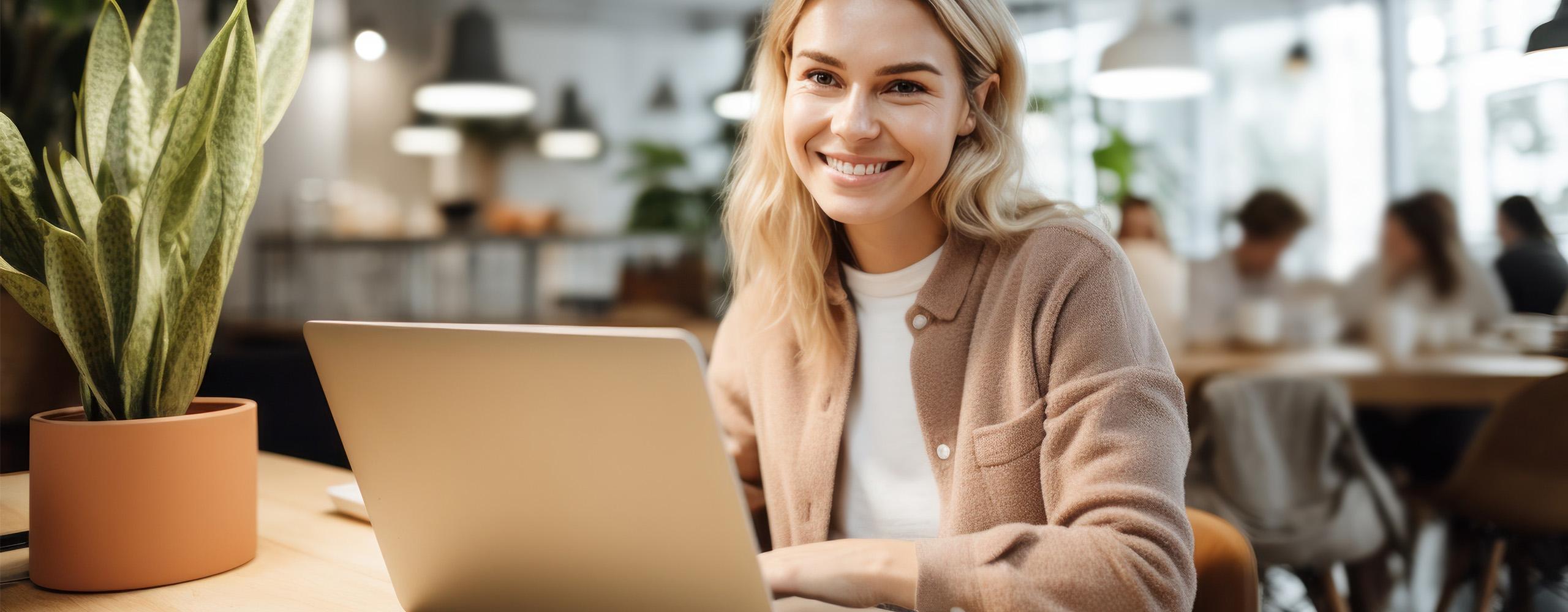 Eine Frau sitzt an einem Tisch vor ihrem aufgeklappten Laptop und lächelt in die Kamera. Im Hintergrund sind weitere Menschen.