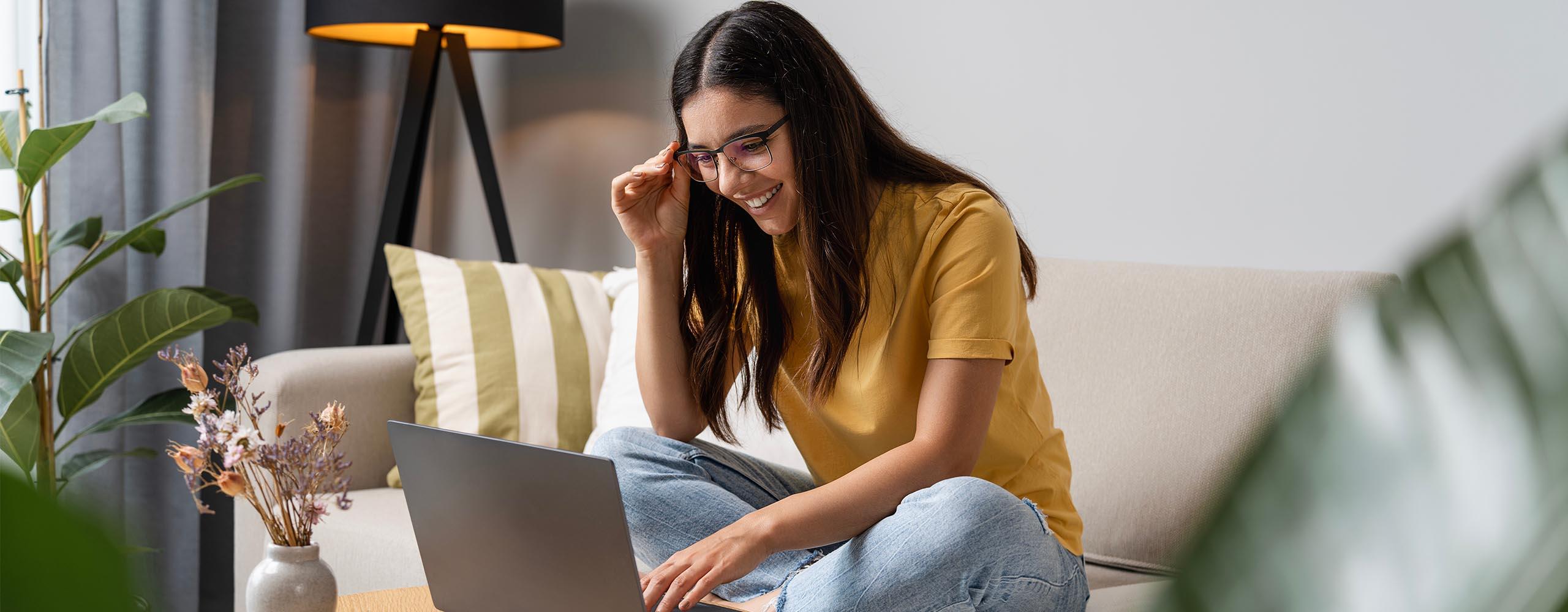 Eine junge Frau sitzt lachend auf dem Sofa vor ihrem Laptop. Mit einer Hand hält sie die Brille in ihrem Gesicht, mit der anderen berührt sie die Tastatur.