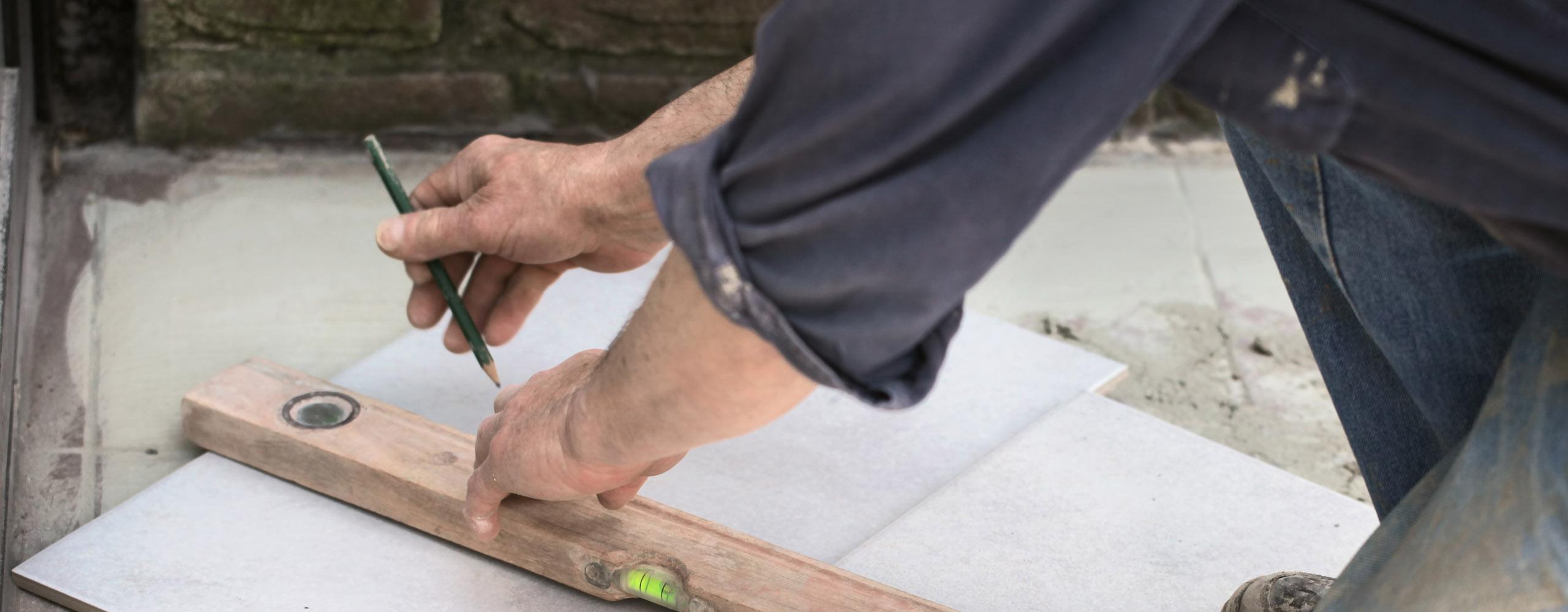 Ein Handwerker zeichnet mit einem Bleistift eine Vermessung auf eine weiße Fliese, damit der Boden nach dem Verlegen keine Unebenheiten aufweist.