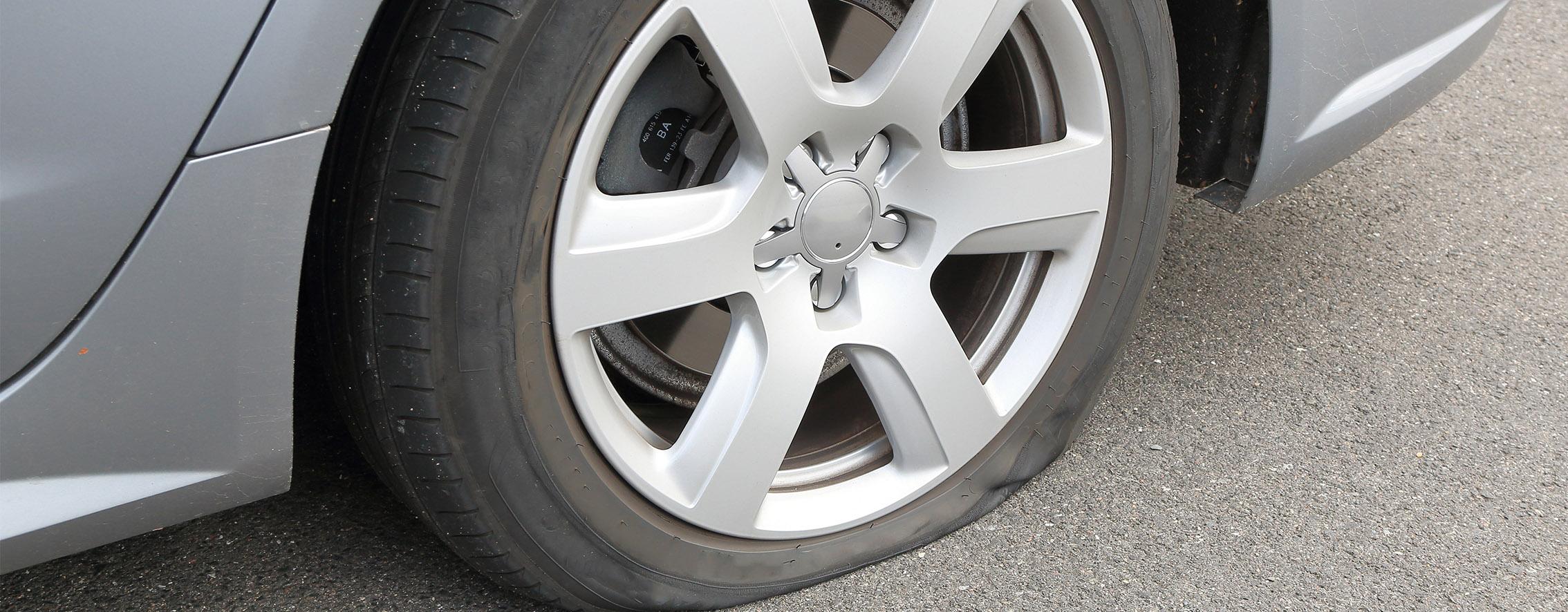 Ansicht auf das linke Hinterrad eines grauen PKWs, das platt ist und für das ein Reifenpannenset benötigt wird