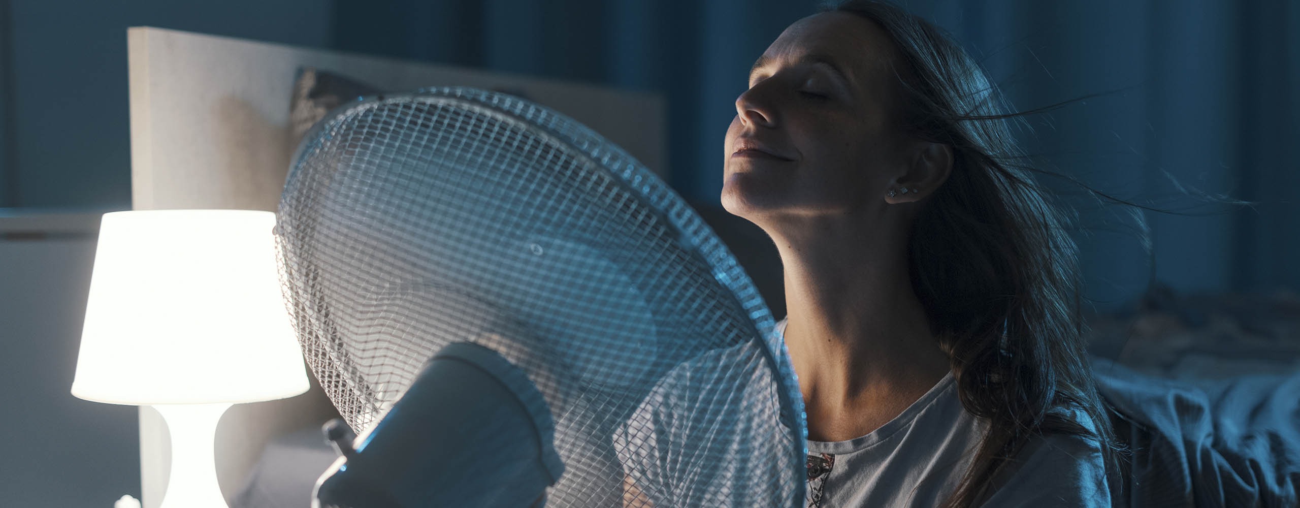 Eine Frau sitzt mit geschlossenen Augen vor ihrem Bett und hält einen eingeschalteten Ventilator vor ihr Gesicht.